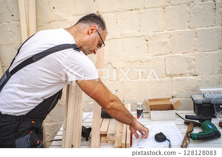 Mature male carpenter using electric screwdriver on a wooden plank outdoors 128128083
