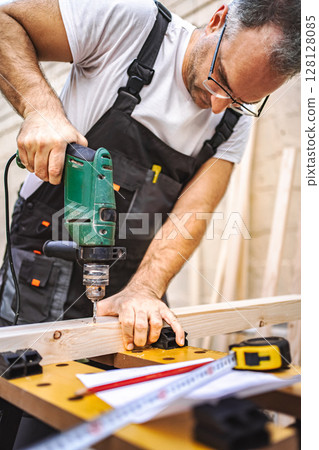 Mature male carpenter using electric screwdriver on a wooden plank outdoors 128128085