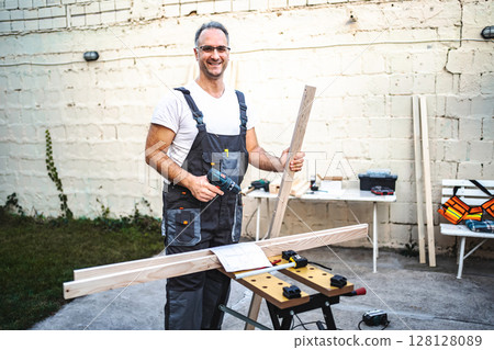 Mature male carpenter working with electrical screwdriver on a wooden plank outdoors 128128089