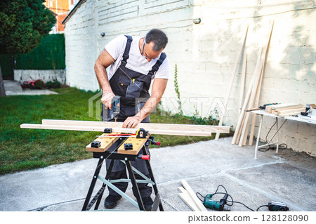 Mature male carpenter using electric drill on a wooden plank outdoors 128128090