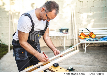 Mature male carpenter measuring wooden plank outdoors 128128091