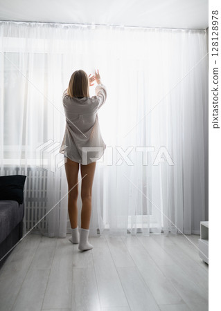 Young woman opening curtains to bright morning light at home. 128128978