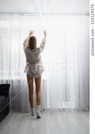 Young woman opening curtains to bright morning light at home. 128128979