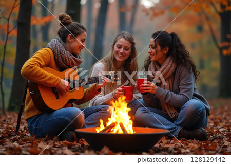 Three women enjoying a campfire together, playing guitar and drinking from red mugs. 128129442