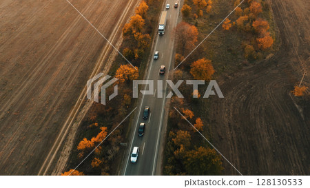 An aerial view of traffic on a road during autumn sunset. Cars driving in a flow of cars along the rural highway surrounded by fields at evening. 128130533