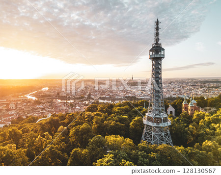 Sunlight breaks over the horizon, illuminating Petrin Lookout Tower and the lush greenery surrounding it. The city of Prague awakens with vibrant hues, creating a stunning panorama. 128130567