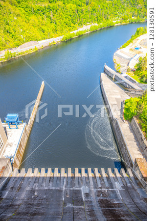 Looking down from the Orlik Dam in Czechia, the calm waters flow beneath, showcasing the engineering marvel of the hydroelectric power station and surrounding lush nature. 128130591