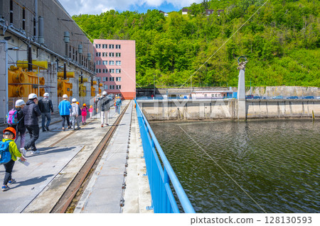 Visitors are seen walking near the Orlik Dam and Hydroelectric Power Station in Czechia. The location features machinery and lush greenery along the riverbank, illustrating energy production. 128130593