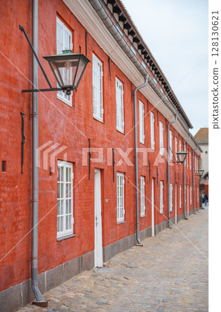Walking along the cobblestone path at Kastellet Barracks in Copenhagen, the vibrant red walls of the historic buildings create a charming atmosphere. Lanterns add to the scenic view. 128130621