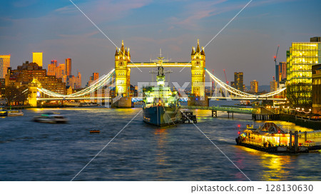 Tower Bridge illuminates in the evening sky, while HMS Belfast rests on the Thames River. Surrounding buildings glow, showcasing London's skyline after sunset. Boats navigate the serene waters. Tower Bridge illuminates in the evening sky, while HMS Belfast rests on the Thames River. Surrounding buildings glow, showcasing London's skyline after sunset. Boats navigate the serene waters. 128130630