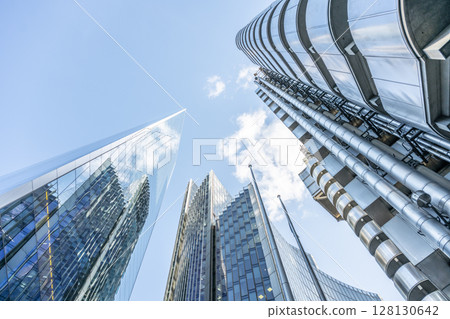 Towering skyscrapers dominate the skyline of London's financial district, showcasing sleek glass facades that reflect the clear blue sky. The urban landscape embodies modern architecture. Towering skyscrapers dominate the skyline of London's financial district, showcasing sleek glass facades that reflect the clear blue sky. The urban landscape embodies modern architecture. 128130642