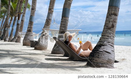 Young woman lounges on sunbed sipping mango shake by ocean on tropical beach 128130664