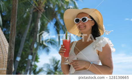 Young woman in swimsuit and sunglasses sips watermelon shake by palm trees on sunny beach 128130665