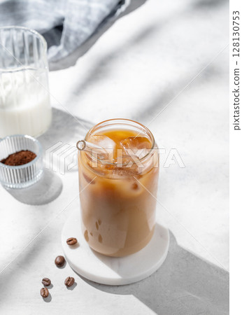 Ice latte coffee in a glass with milk on a light background with coffee beans and morning shadows. Ice latte coffee in a glass with milk on a light background with coffee beans and morning shadows. 128130753