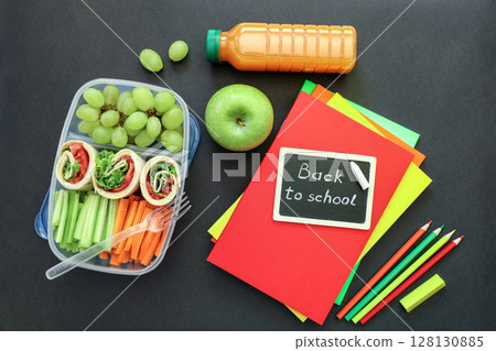 School supplies and lunch box with tasty rolls, cucumbers, carrots, grapes, apple, bottle of juice on black background, back to school concept. Horizontal. Top view. 128130885