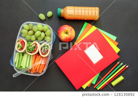 Box contains wraps with vegetables, grapes, apple, juice bottle, colorful notebooks and pencils arranged neatly on black background for school lunch preparation. 128130892