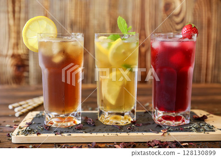 Three tall glasses filled with iced tea, lemonade with lemon slices, and a berry drink with cherries are presented on rustic wooden table. Fresh mint garnish adds vibrant touch. 128130899