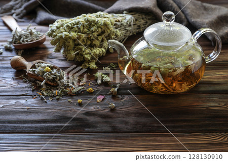Tea is steeping in glass teapot, surrounded by fresh herbs and dried flowers on weathered wooden table, creating warm and inviting atmosphere for relaxation and enjoyment. Tea is steeping in glass teapot, surrounded by fresh herbs and dried flowers on weathered wooden table, creating warm and inviting atmosphere for relaxation and enjoyment. 128130910