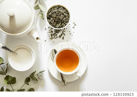 Gentle steam rises from freshly brewed tea in white cup beside herbal leaves in bowl and sugar in dish, surrounded by eucalyptus branches on bright white background. Gentle steam rises from freshly brewed tea in white cup beside herbal leaves in bowl and sugar in dish, surrounded by eucalyptus branches on bright white background. 128130923