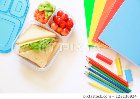 Lunch box filled with sandwiches, cucumber, carrots and cherries placed next to vibrant stationery, including notebooks and colored pencils, ideal for school activities on white backgrond. Lunch box filled with sandwiches, cucumber, carrots and cherries placed next to vibrant stationery, including notebooks and colored pencils, ideal for school activities on white backgrond. 128130924
