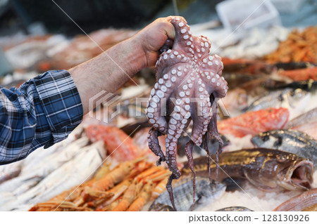 Vendor showcases freshly caught octopus at seafood market in Greece. Various types of fish and shellfish surround vendor, creating a lively atmosphere. Horizontal. 128130926