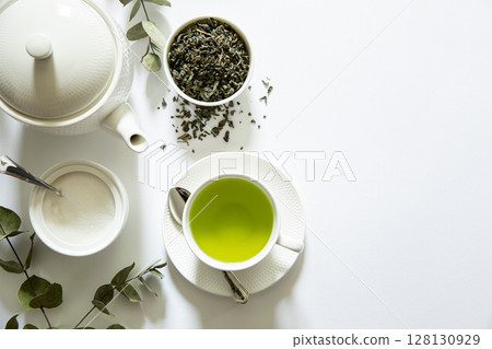 Bright green tea prepared in a simple cup alongside loose tea leaves, sugar bowl, and eucalyptus branches creating a calming atmosphere in a minimalist setting. 128130929