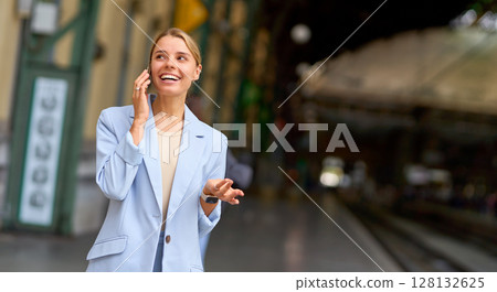 A happy businesswoman talks on her phone at a busy train station, reflecting her active career 128132625