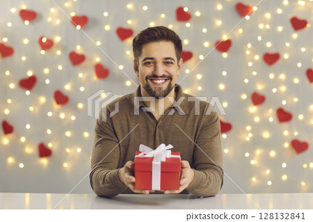 Smiling man boyfriend sitting at table, holding out holiday present box and looking at camera 128132841
