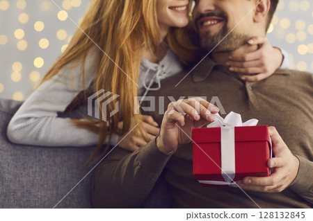 Smiling woman embracing her boyfriend who's opening his Valentine or birthday present Smiling woman embracing her boyfriend who's opening his Valentine or birthday present 128132845