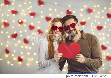 Young happy loving couple in heart shaped glasses standing and holding big red heart in hands Young happy loving couple in heart shaped glasses standing and holding big red heart in hands 128132846