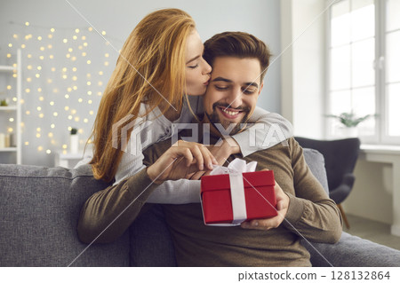 Young redhead wife hugs and kisses her husband on the cheek giving him a gift for Valentine's Day. 128132864