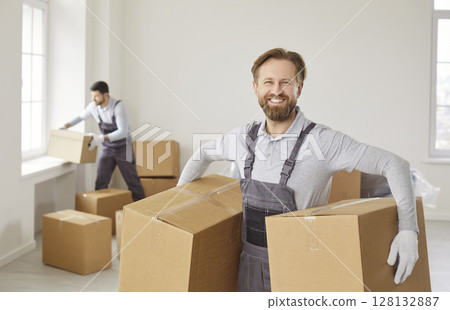 Smiling mover holding boxes in empty room, assisting in relocation process in new apartment. Smiling mover holding boxes in empty room, assisting in relocation process in new apartment. 128132887
