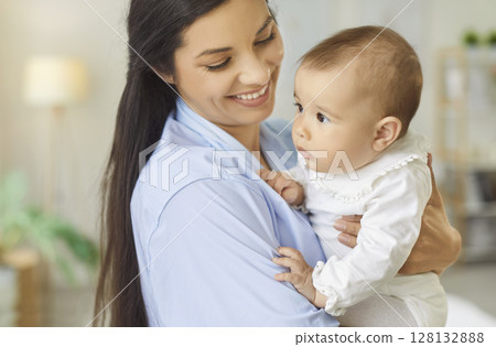 Closeup portrait of smiling happy young mother holding little cute baby in hands standing at home Closeup portrait of smiling happy young mother holding little cute baby in hands standing at home 128132888