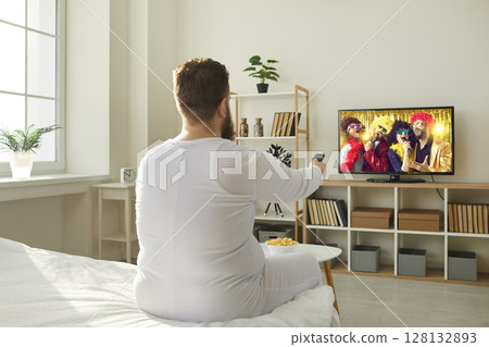 Funny chubby guy in pajamas sitting in bedroom and watching TV show on television Funny chubby guy in pajamas sitting in bedroom and watching TV show on television 128132893