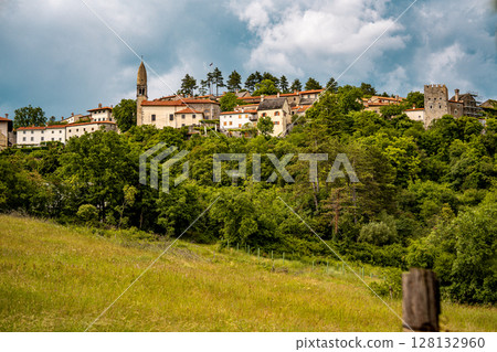 Scenic Hilltop Town Of Stanjel In Slovenia: Traditional Stone Houses And Historic Church Tower Rising Above Lush Greenery Under A Dramatic Cloudy Sky In The Karst Region 128132960