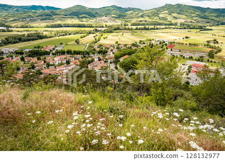 Picturesque View Of Vipava From Castle Ruins: Red-Tiled Rooftops And Church Tower Nestled In Green Valley With Blooming Flowers In Foreground And Rolling Fields In Background, Slovenia 128132977