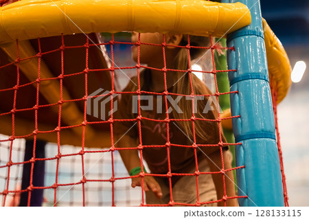 A little girl happily explores a colorful playground, holding on to a red safety net and moving around the play area in an amusement park. A child climbs through a maze in a playroom. 128133115