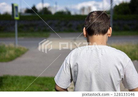 Young boy wearing a white t-shirt standing near a pedestrian crossing on a bright, sunny summer day, contemplating the decision to cross the street while enjoying the warmth of the outdoors. 128133146
