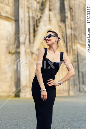 Portrait young adult elegant woman black dress and sunglasses smiles confidently in golden sunlight warm sunset backlit light against historic cathedral building. Female person walking city street Portrait young adult elegant woman black dress and sunglasses smiles confidently in golden sunlight warm sunset backlit light against historic cathedral building. Female person walking city street 128133574