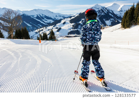 Scenic portrait view kid beginner skier close-up downhill start line rows freshly prepared groomed ski slope piste bright warm morning sunrise sky background.  mountain landscape winter resort 128133577