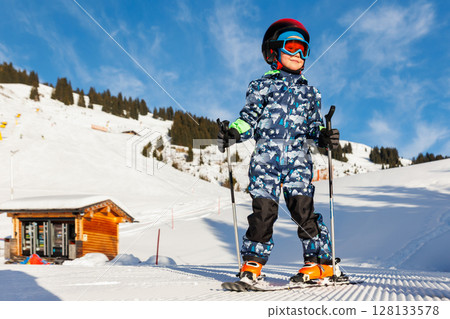 Scenic portrait view kid beginner skier close-up downhill start line rows freshly prepared groomed ski slope piste bright warm morning sunrise sky background. mountain landscape winter resort Scenic portrait view kid beginner skier close-up downhill start line rows freshly prepared groomed ski slope piste bright warm morning sunrise sky background. mountain landscape winter resort 128133578