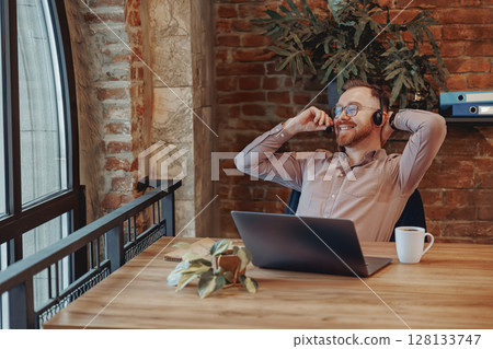 A Relaxed Professional in a Modern Workspace Utilizing a Laptop and Wearing Headphones 128133747