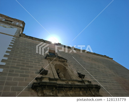 As the sun rises, golden rays light up the historical church and the statue of the saint in Denia, creating a serene morning atmosphere. Visitors can enjoy the peaceful setting. As the sun rises, golden rays light up the historical church and the statue of the saint in Denia, creating a serene morning atmosphere. Visitors can enjoy the peaceful setting. 128133779