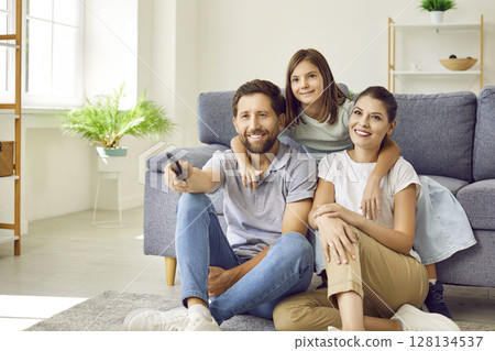 Happy mom, dad and child sitting on floor by sofa and watching television show together Happy mom, dad and child sitting on floor by sofa and watching television show together 128134537
