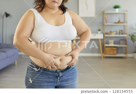 Fat overweight young woman sadly touching her big belly standing in living room at home. Fat overweight young woman sadly touching her big belly standing in living room at home. 128134555