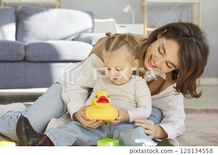 Smiling mother and little daughter playing with yellow rubber duck on floor while bonding at home. 128134558