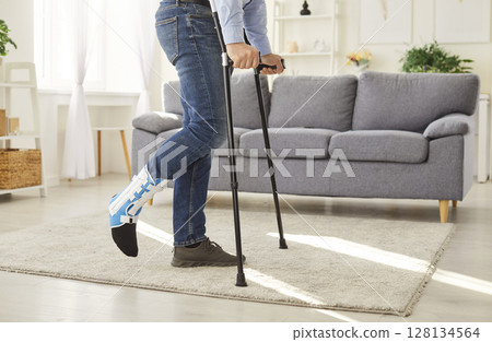 Young man with broken leg after accident using crutches standing at home with plastered leg. 128134564