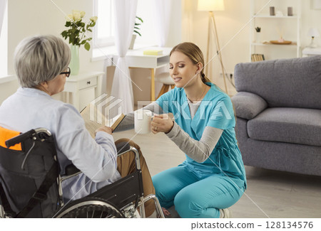 Young caregiver giving senior mature woman with disability in wheelchair a cup of beverage. 128134576