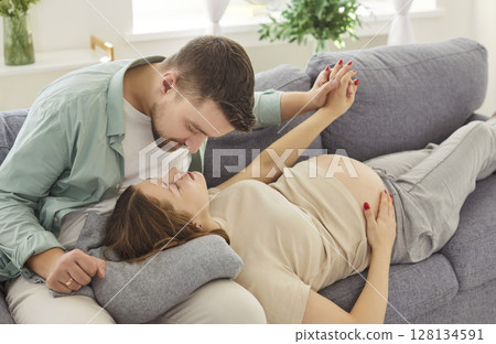 Young man sitting on sofa at home with pregnant wife looking at each other with tenderness Young man sitting on sofa at home with pregnant wife looking at each other with tenderness 128134591