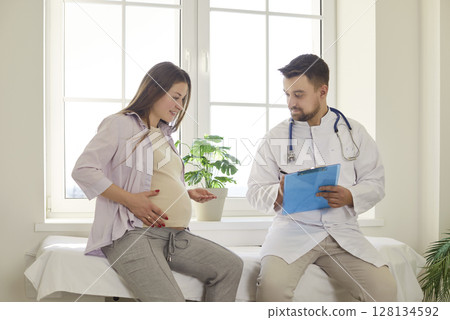 Pregnant woman talking to male doctor during consultation and prenatal checkup in medical office. 128134592